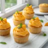 golden mango cupcakes with swirled orange buttercream frosting on a white marble surface